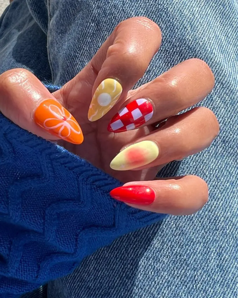 A hand with a summer-themed manicure featuring orange with white flower designs, yellow with a daisy, and a red and white gingham pattern, contrasted against blue fabric and denim.