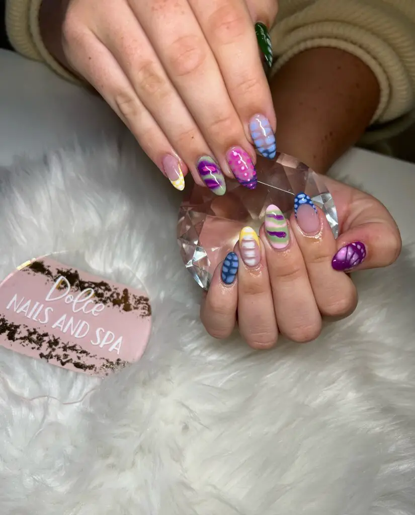 A hand with nails painted in various patterns of lavender and green, some with a matte finish and others glossy, showcasing an intricate mix of swirls and stripes.