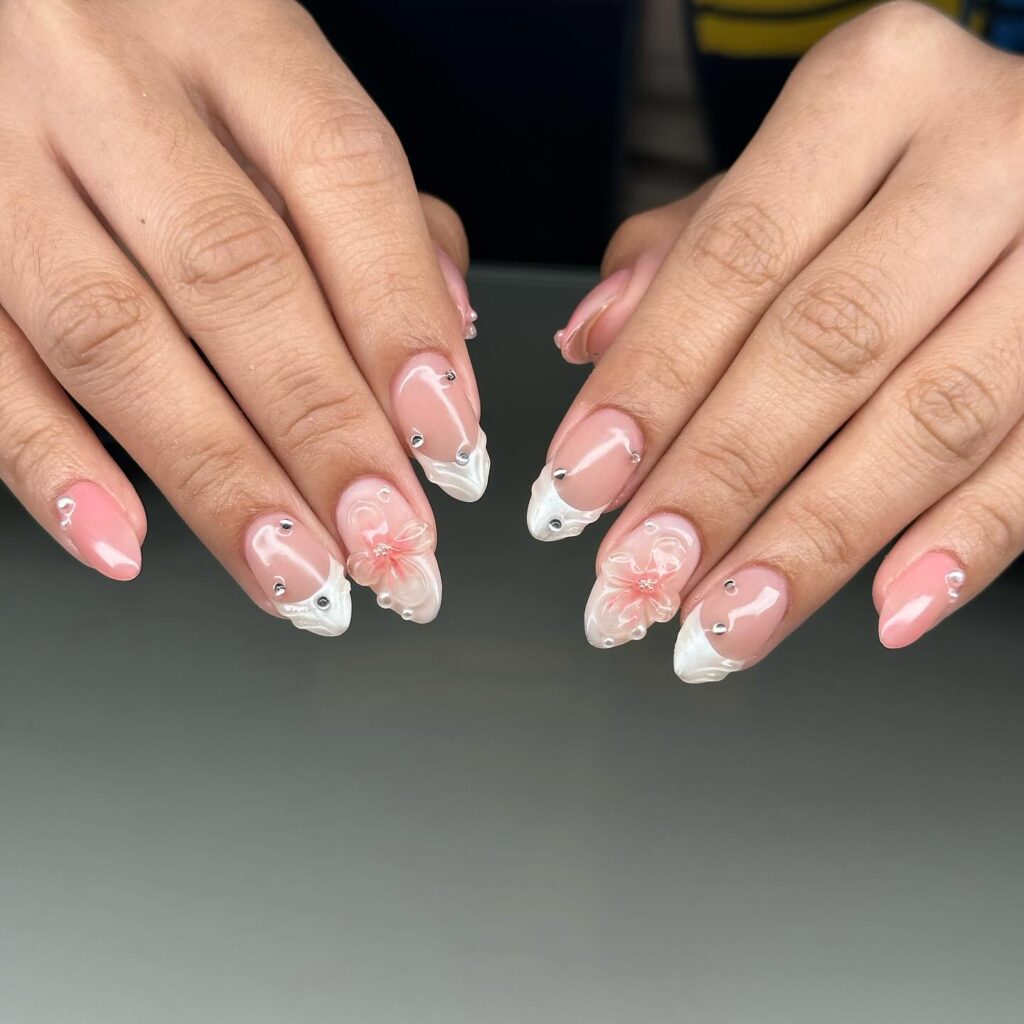 A hand adorned with long nails featuring a sheer pink base and white 3D floral designs with rhinestone centers for an elegant touch.