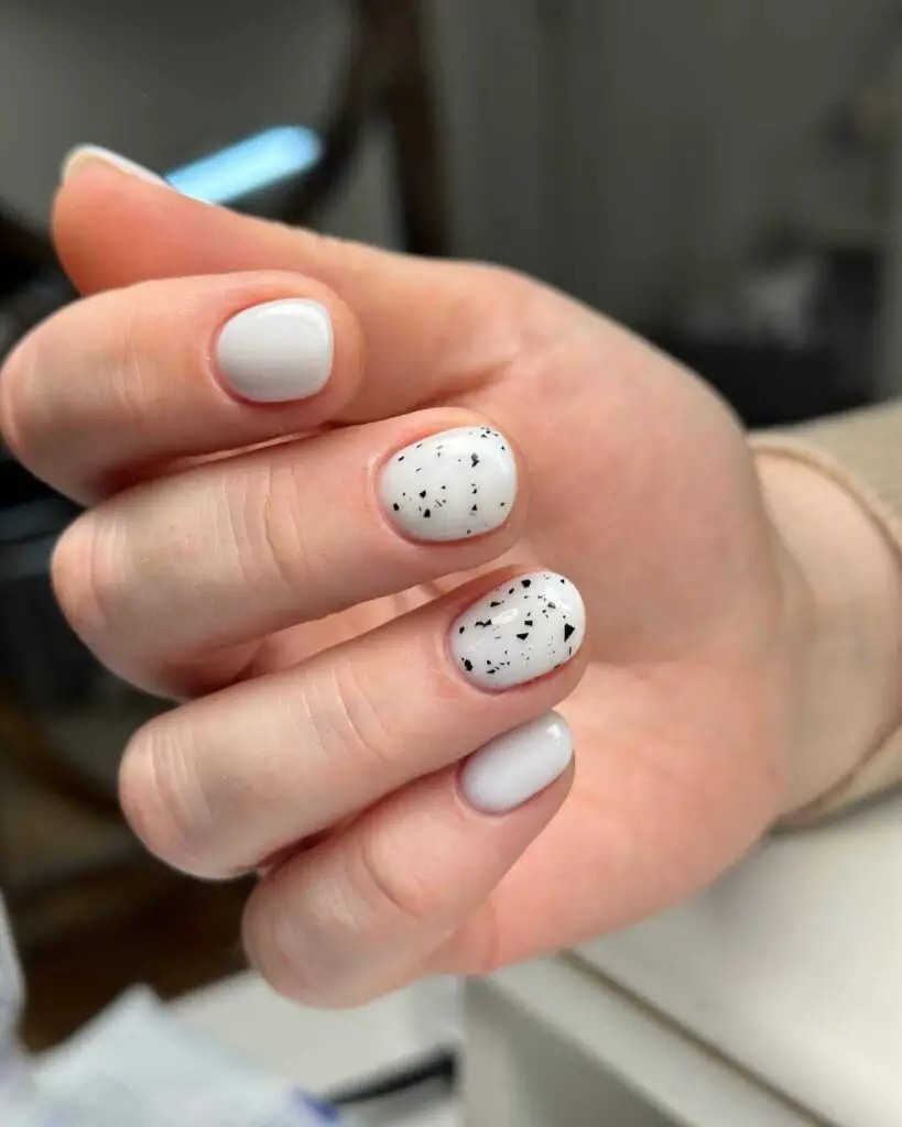 Short nails painted white with black speckles on two fingers, displaying a clean and minimalist design.