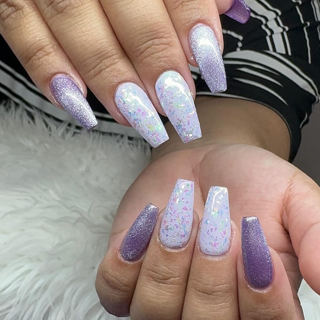 A hand with nails painted in alternating shades of lavender and white confetti-patterned polish, evoking a light and playful style.