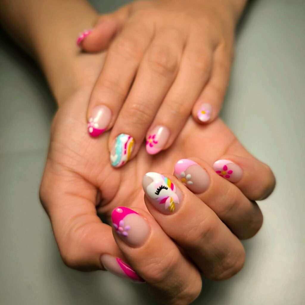 A display of creative nail art with designs including a unicorn, rainbows, and bright pink accents on a pastel base, exuding a playful and artistic vibe.