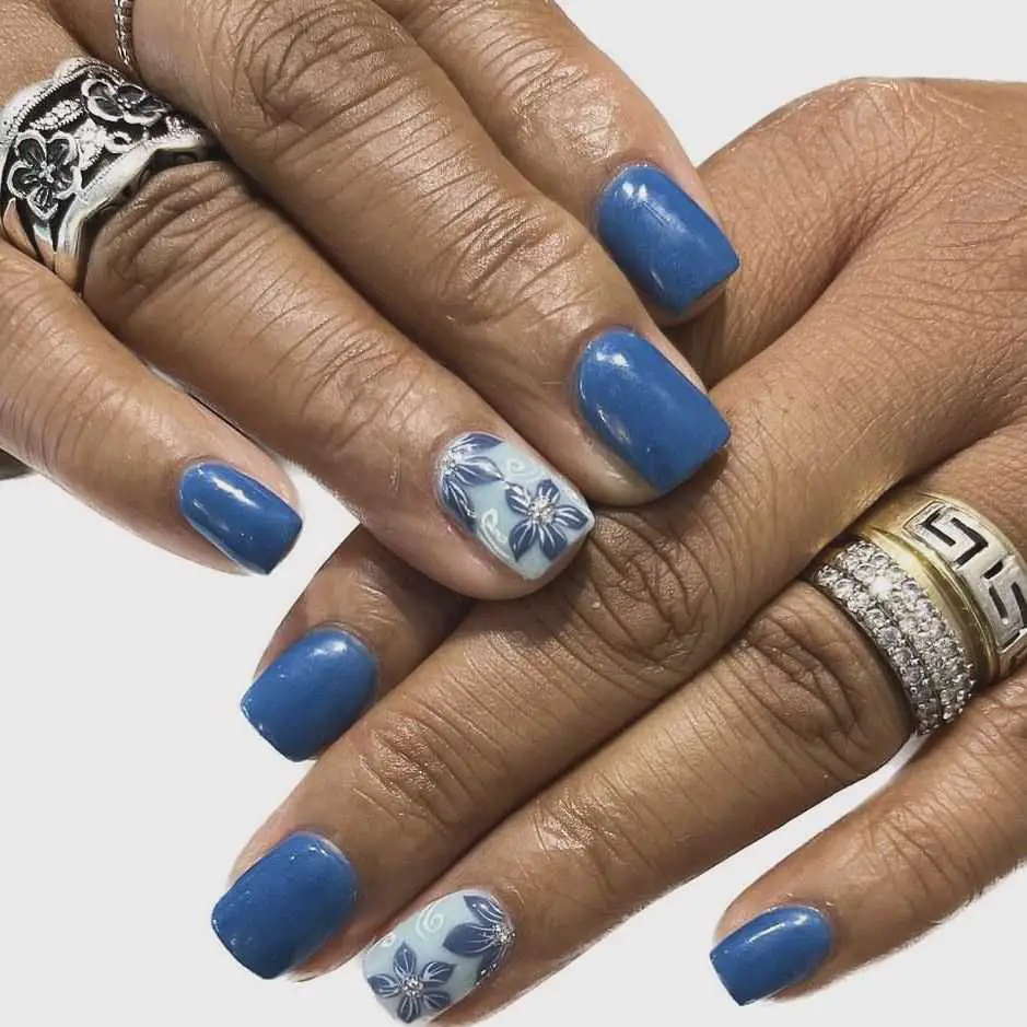 Hands with nails painted in a striking cobalt blue adorned with white floral details, exemplifying a bold yet elegant look.