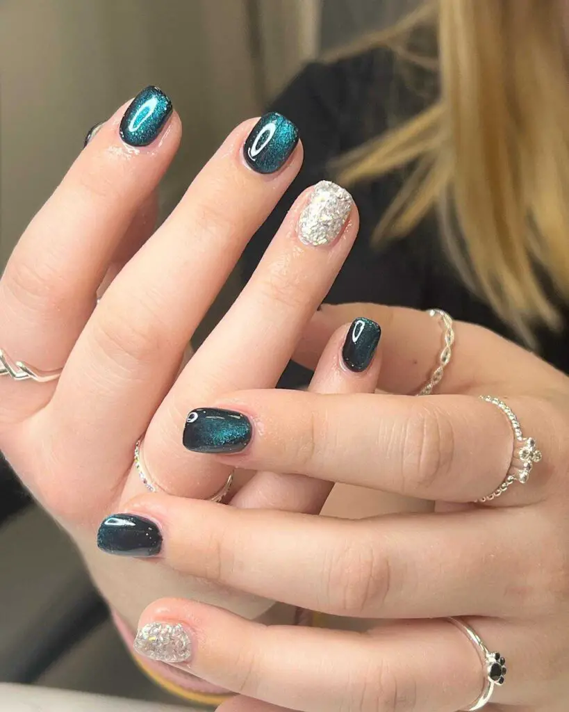 Close-up of a hand with nails painted in dark glossy tones with one accent nail featuring sparkling silver flecks, inspired by a starry night sky.