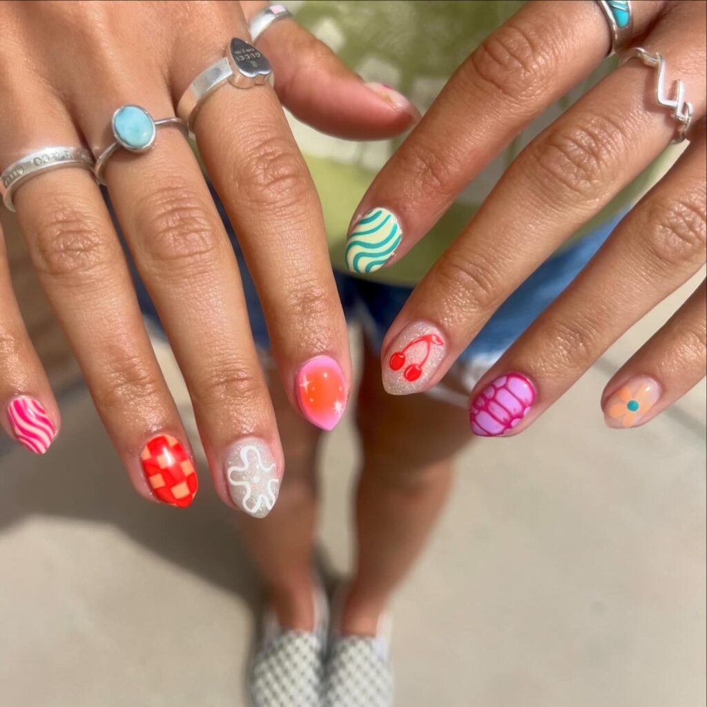 A hand showcasing a mix of playful summer-themed nail designs including ocean waves, strawberries, and pastel patterns, perfect for an adventurous spirit.