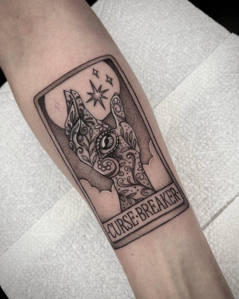 An arm tattoo designed like a 'Curse Breaker' tarot card with mystical symbols, suggesting overcoming and insight.