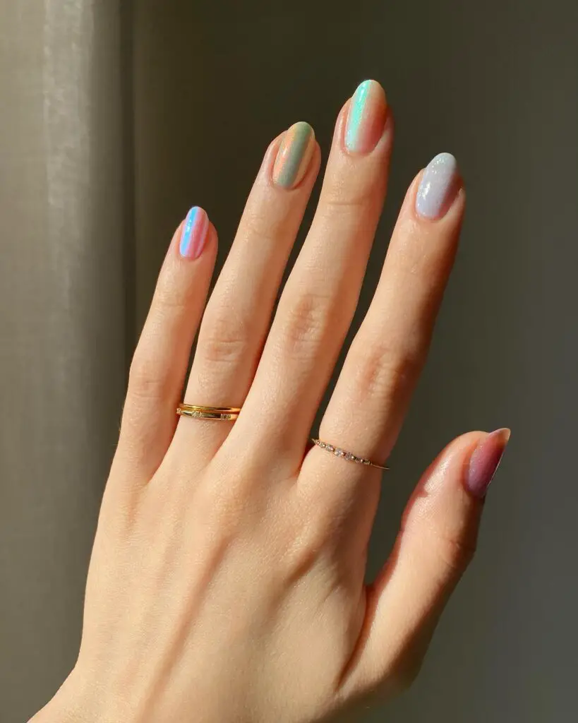 Hand with nails painted in pastel iridescent colors creating a holographic rainbow effect, accompanied by delicate gold rings.