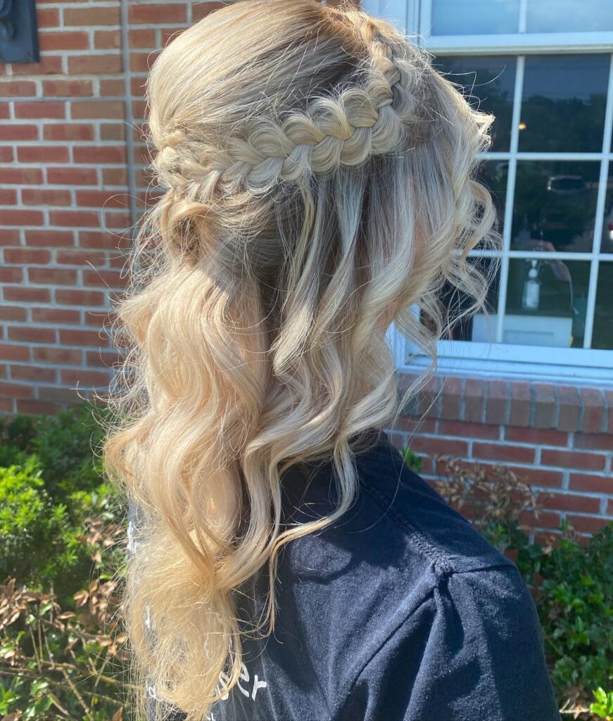 A woman's blonde hair styled with a crown braid and loose waves, capturing the warm glow of sunlight for a radiant prom look.