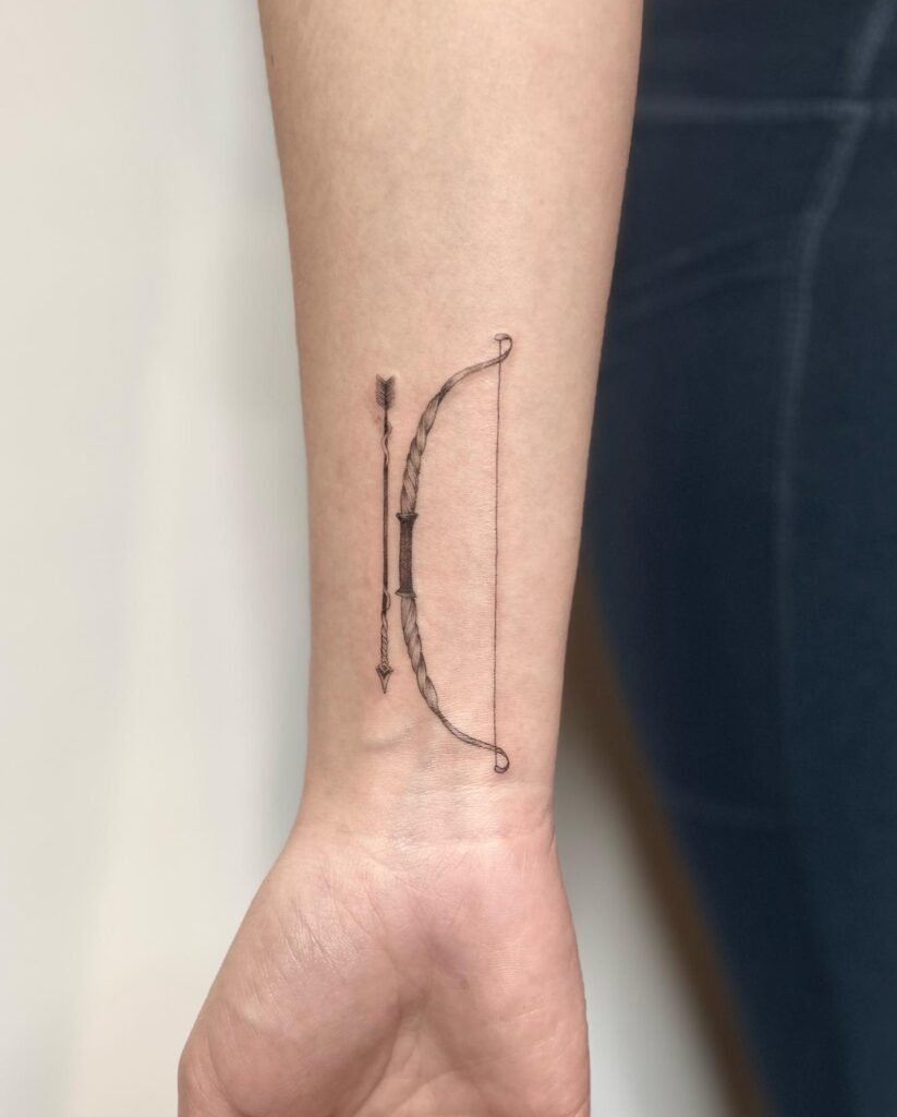 A simple wrist tattoo of a bow and arrow, signifying focus, direction, and pursuit.