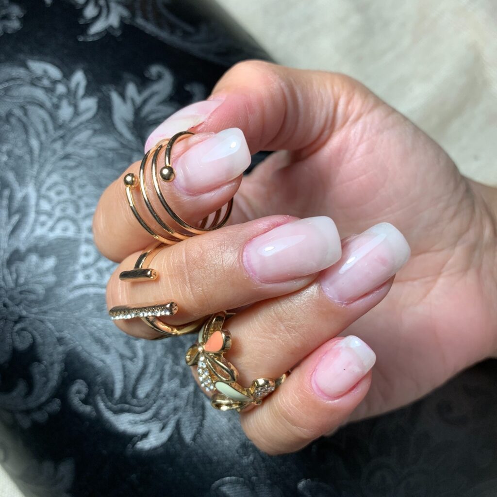 Hands elegantly displaying French ombre nails, transitioning from a natural pink base to white tips, complemented by an array of gold rings.