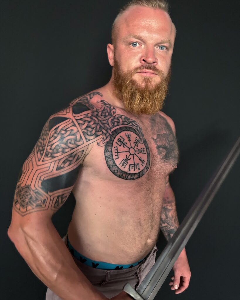 A man with a Vegvísir rune chest tattoo and a Norse mythological scene on his shoulder.