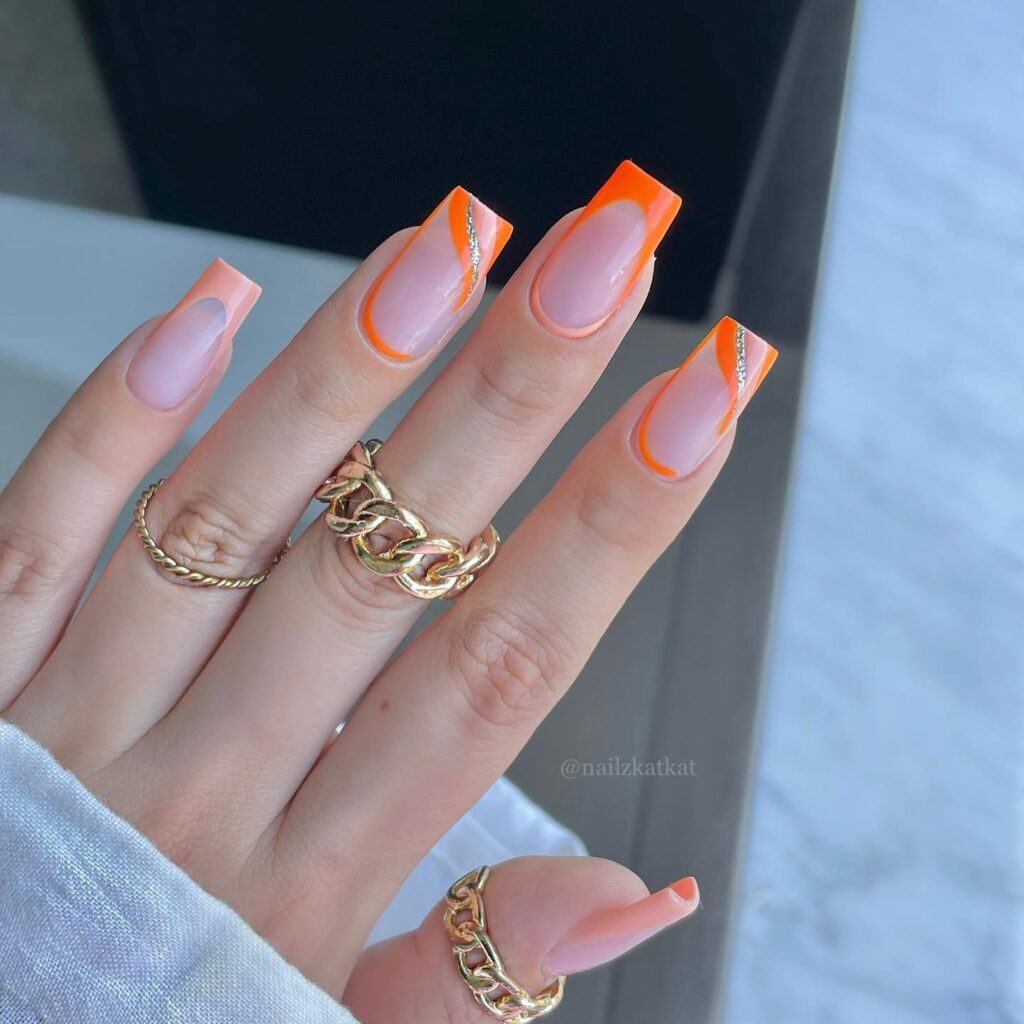 Close-up of a hand with long nails featuring a nude base and vibrant orange French tips, complemented by gold rings.