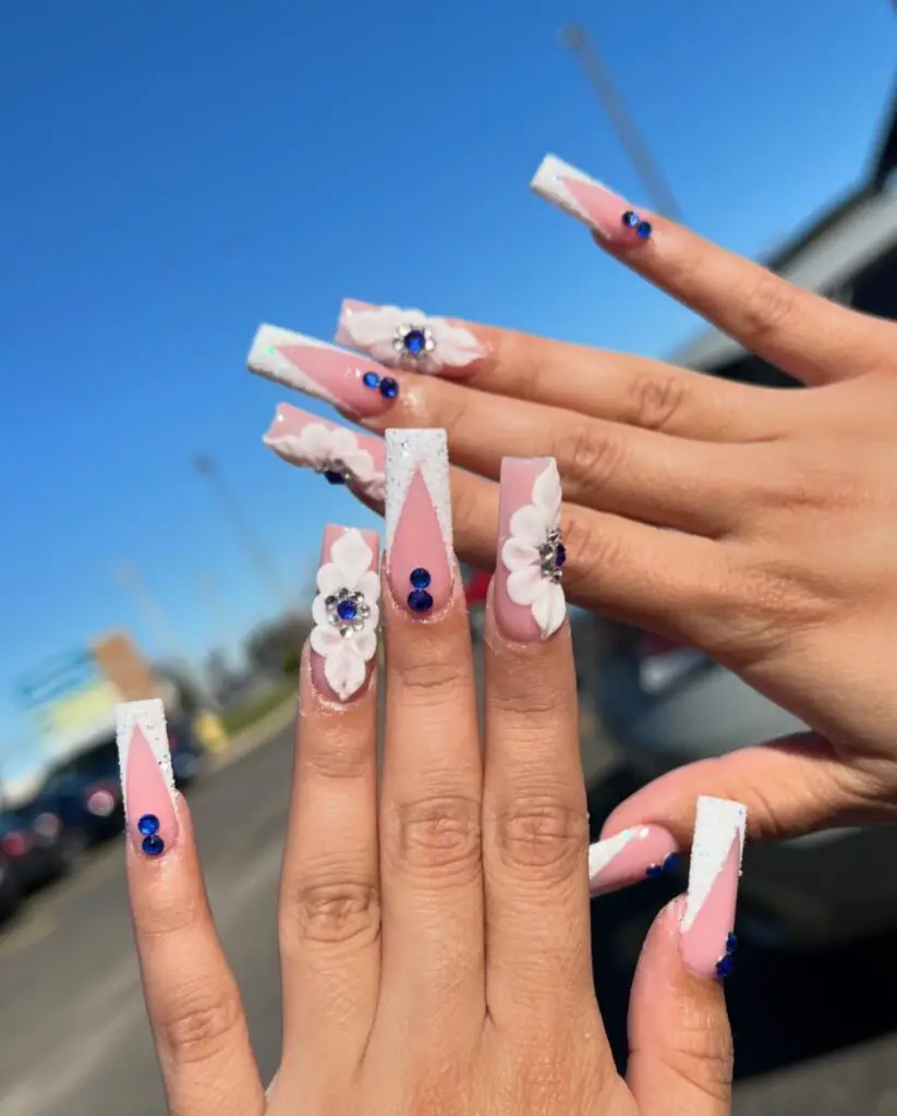 Hands displaying long coffin nails with a soft pink base, glittery diamond dust, blue gemstone embellishments, and white floral appliqués, set against a blue sky background.