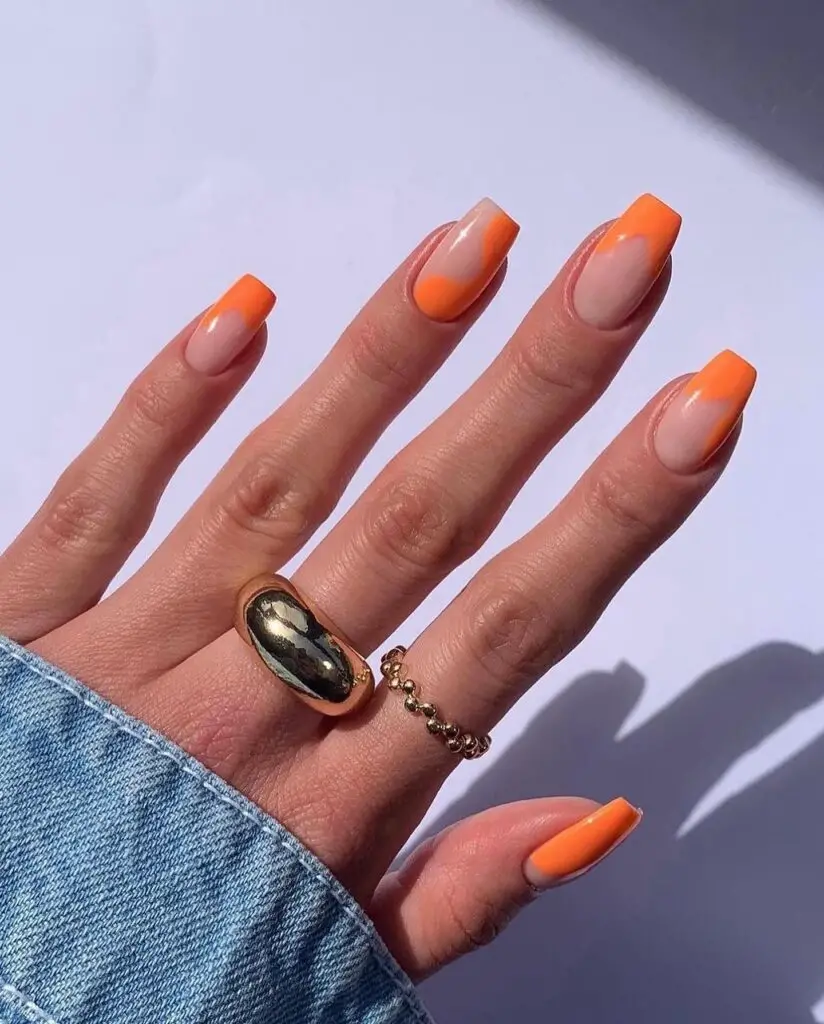 Hand with summer-inspired full-coverage orange nail polish on squared nails, accented by a denim sleeve and a large oval ring.