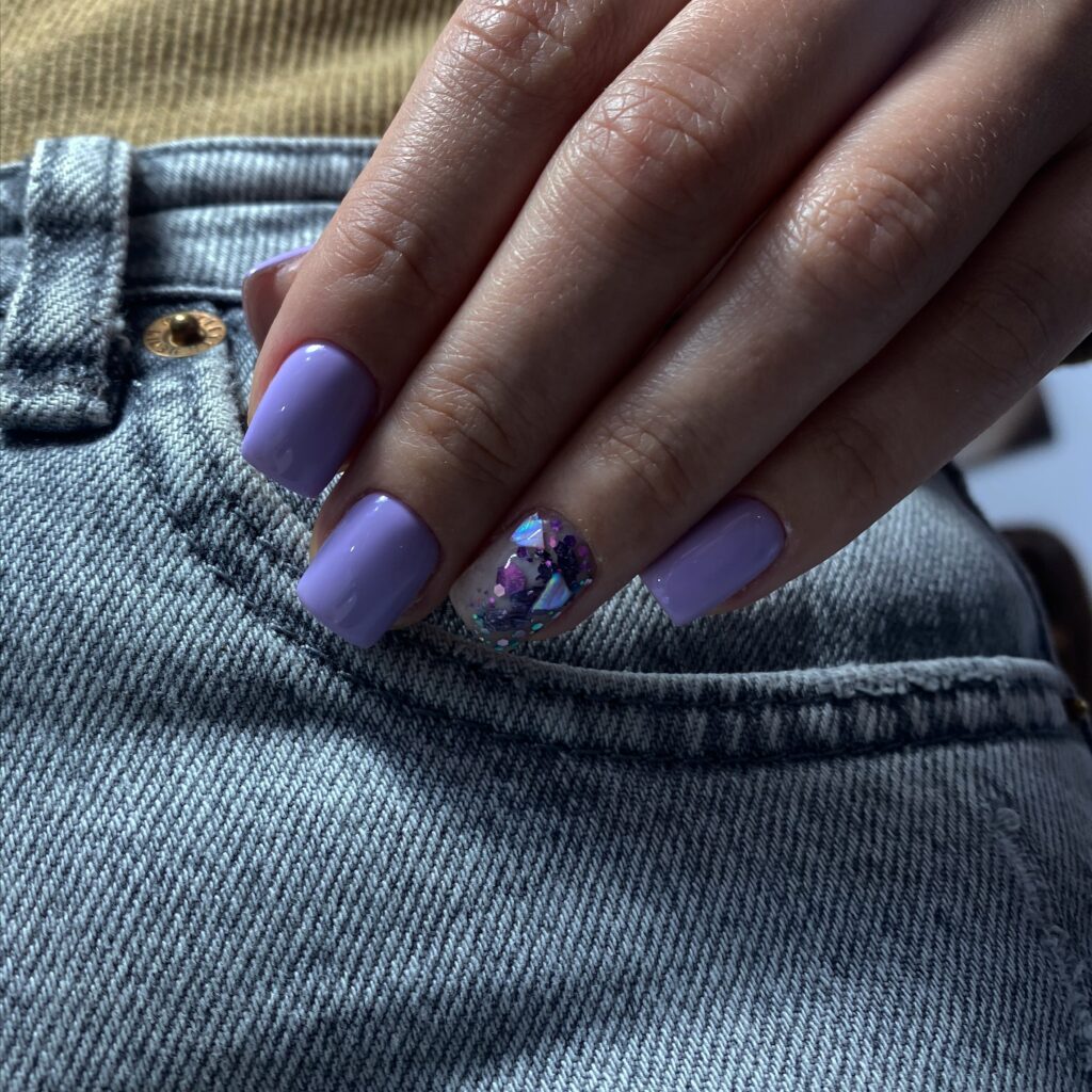Hand with purple painted nails resting on denim fabric, with one nail showcasing a clear polish with purple and blue glitter pieces.