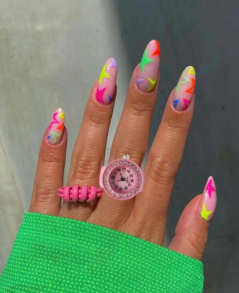 A hand with almond-shaped nails featuring vibrant abstract neon swirls in pink, green, orange, and yellow, paired with a stylish pink ring and green sleeve.