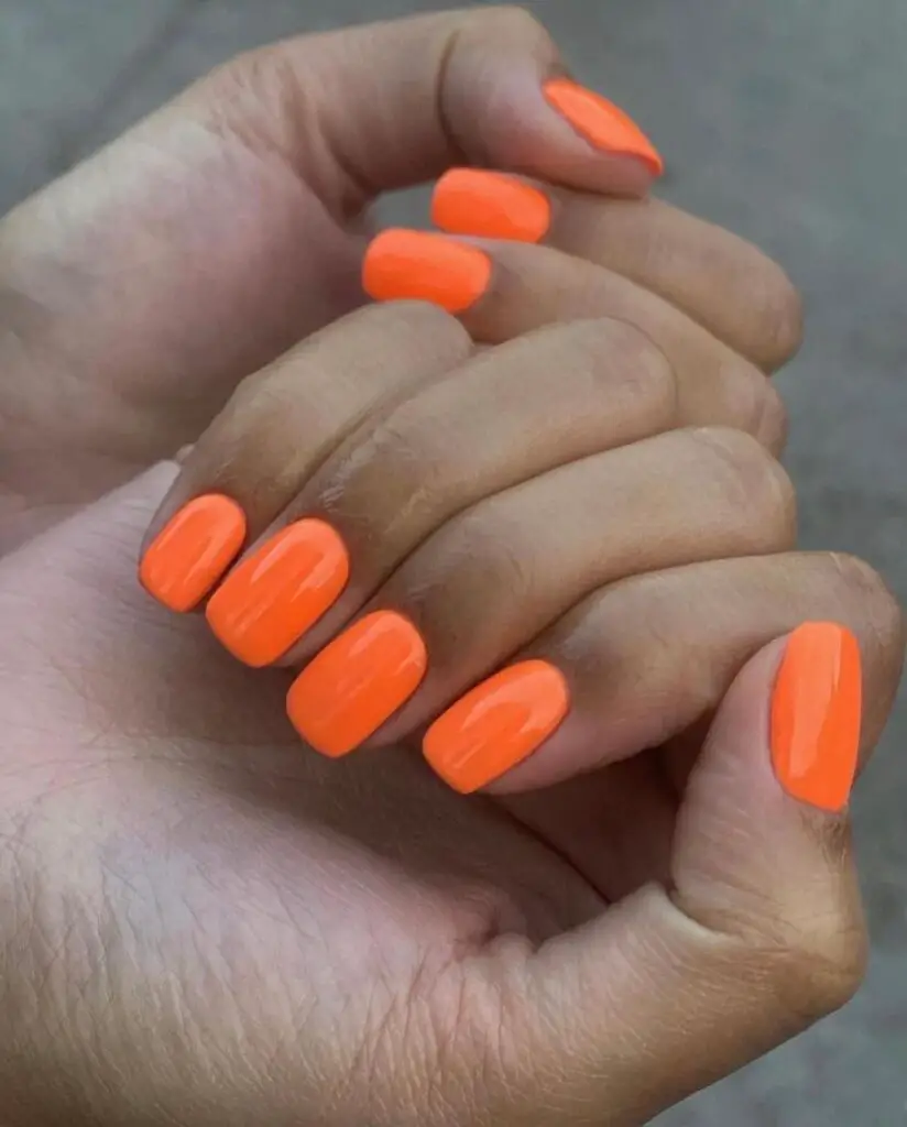 Hand displaying short nails painted with a vivid neon orange polish, perfect for a bold summer statement.
