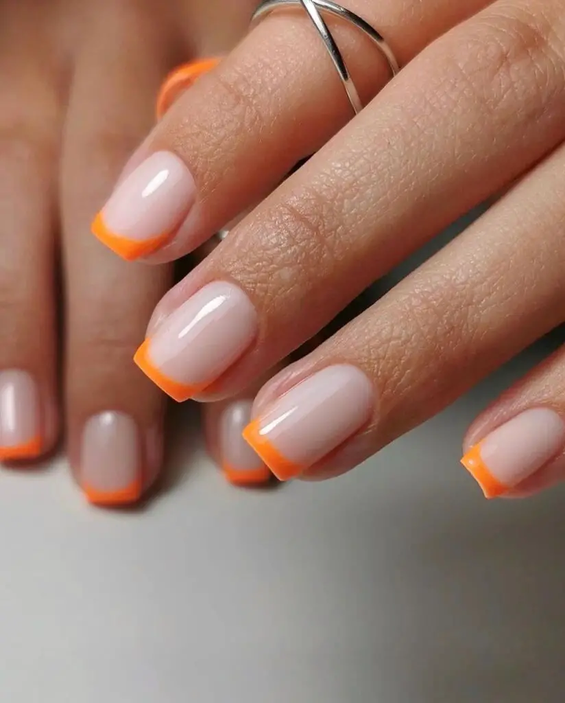 Elegant hand showing off a minimalist nail design with a sheer nude base and bright orange borders on a set of oval nails.