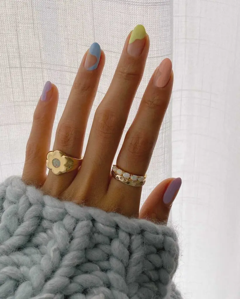 A hand partially covered with a gray knitted sleeve showcases nails with a pastel swirl design in lavender, sky blue, and pale yellow.