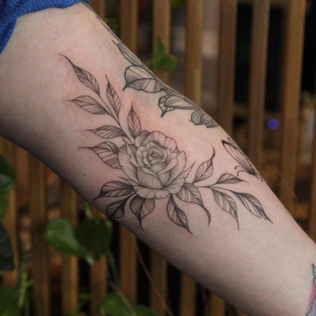 An arm tattoo of a blooming rose with leaves, symbolizing love and full embrace of passion.