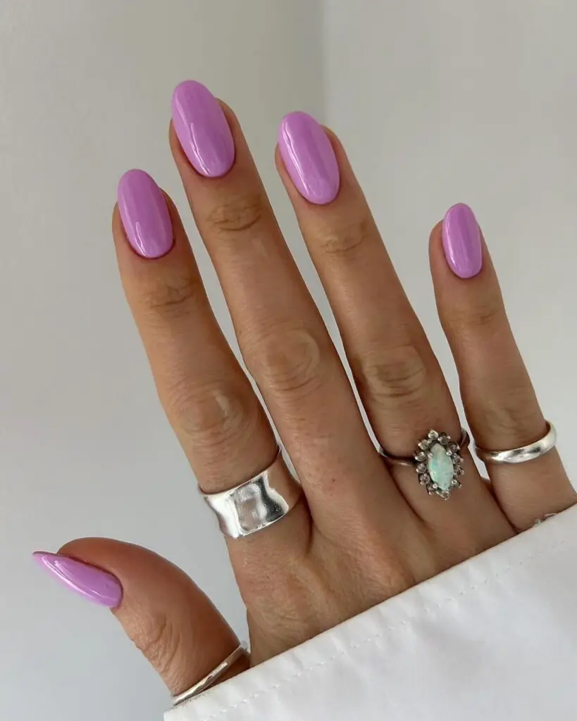 Hand displaying glossy, almond-shaped nails painted in a uniform lavender shade, complemented by stylish silver rings.
