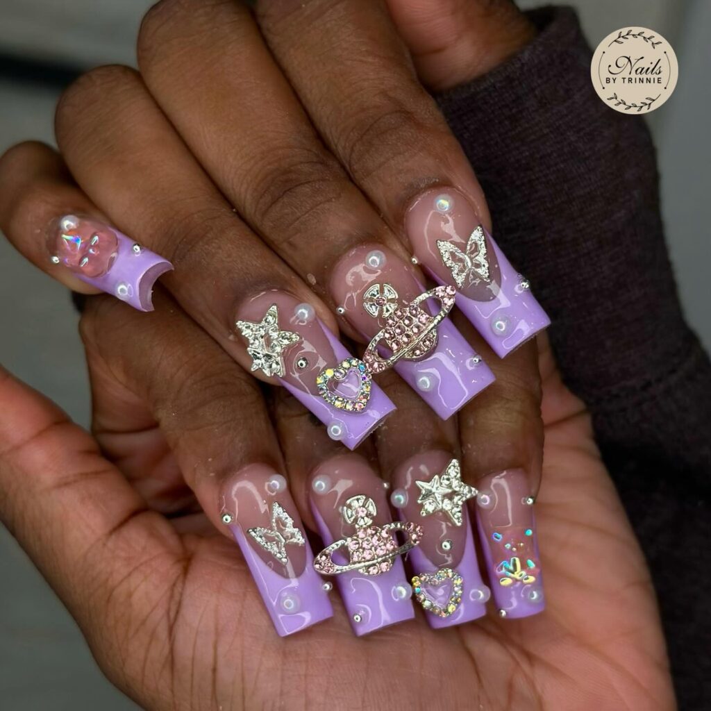 Hands displaying extended translucent lavender nails adorned with intricate rhinestone and metallic decorations, exuding opulence and style.
