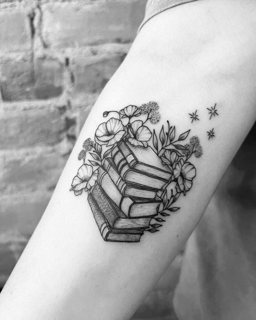 A tattoo of a stack of books intertwined with flowers and falling stars on an arm, symbolizing a love for literature and nature.