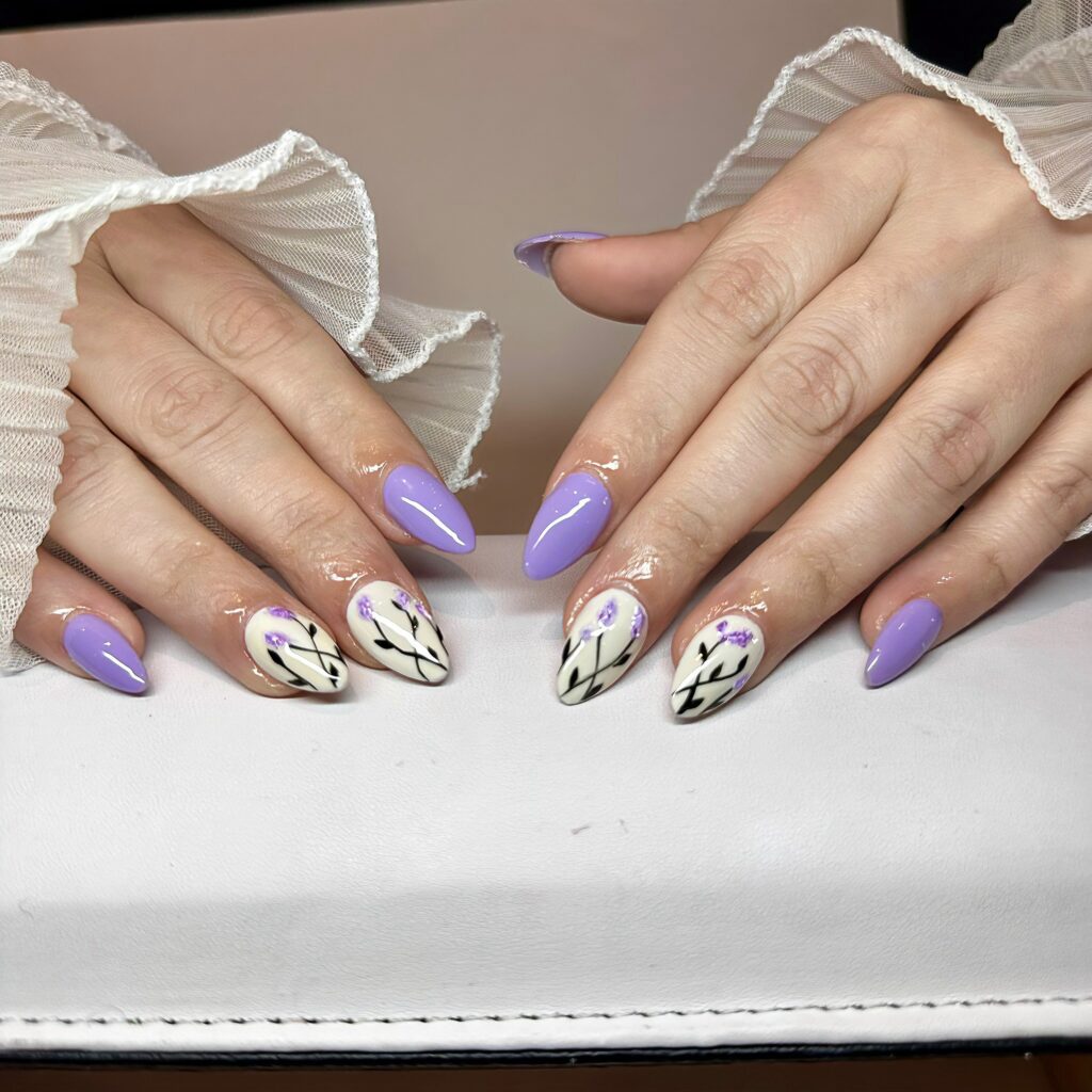 A hand with lavender nails, some adorned with delicate white floral and geometric line art, exemplifying detailed nail artistry.