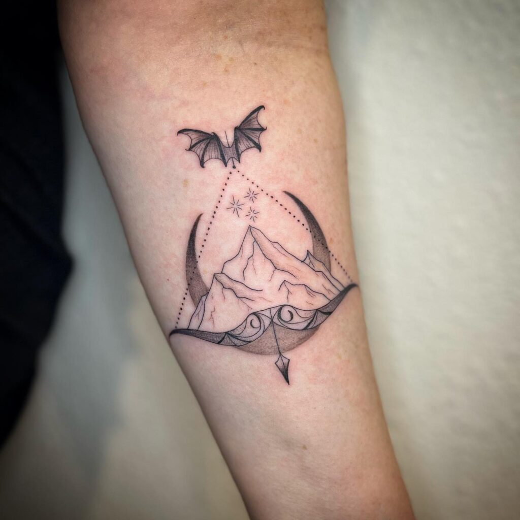 A tattoo of a mountain peak with a bat and celestial elements in a triangular frame on a forearm, representing adventure and the night.