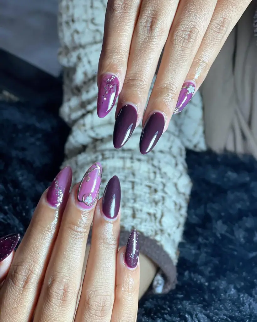 A hand featuring almond-shaped nails in a deep plum color with artistic white splatter and gold fleck accents.