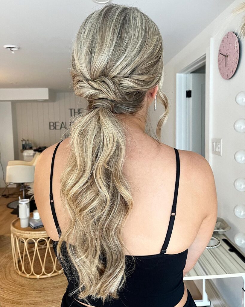 A polished blonde hairstyle with a low twisted ponytail and wavy ends, suited for a sleek prom look.