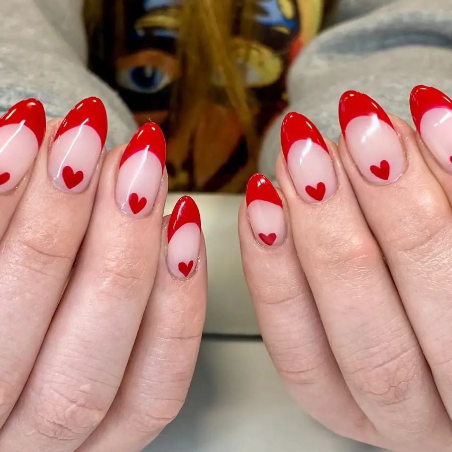Hands with stiletto-shaped nails showcasing a romantic design with red tips and small red hearts on a sheer white base.