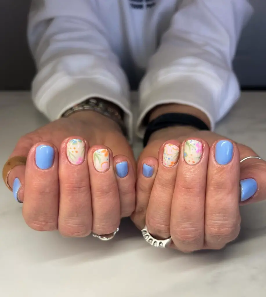 Hands with a mix of solid pastel blue nails and white nails with multicolored floral and abstract patterns, reminiscent of a spring day.