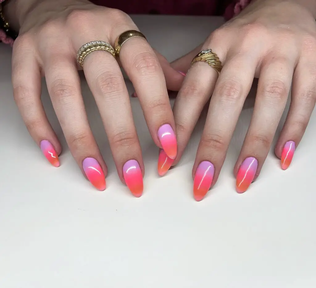 A set of nails featuring a pink to orange ombre effect, resembling a tropical sunset, showcased on a person's hands with gold rings.
