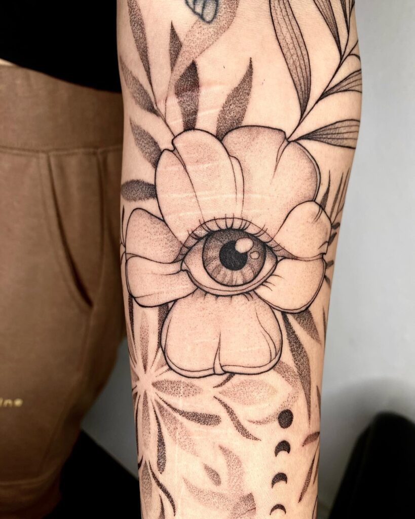 An arm tattoo featuring an eye within a blooming flower, framed by leaves, denoting growth and awareness.