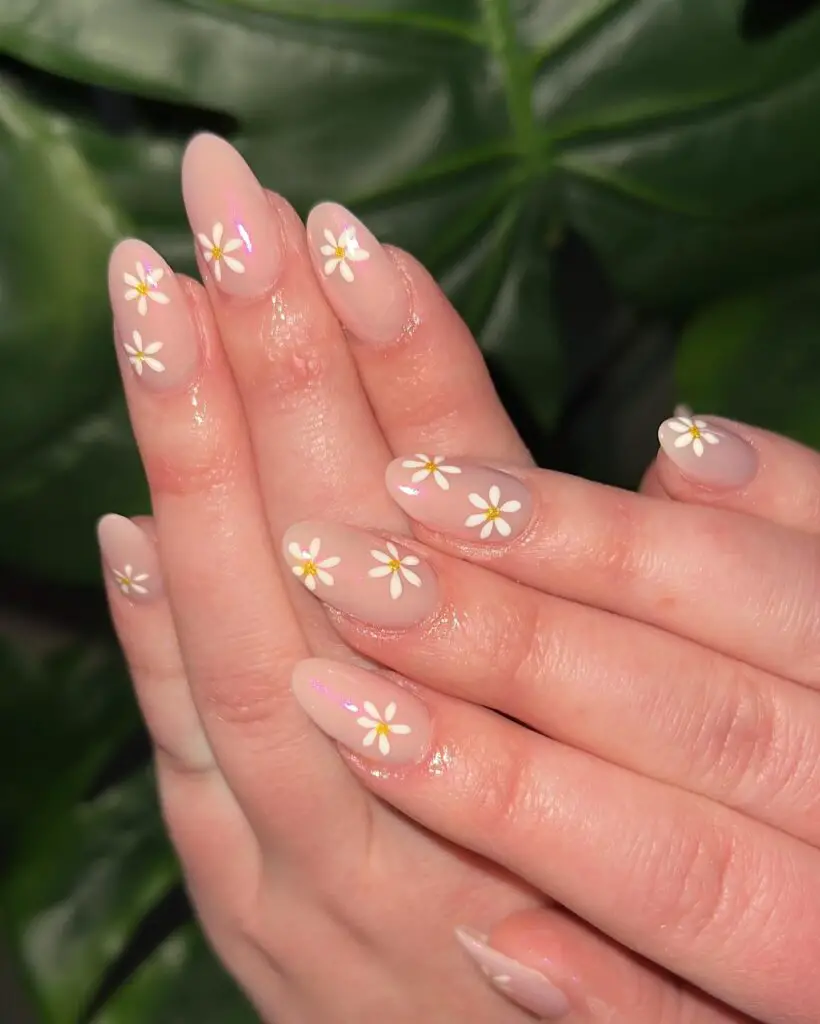 Close-up of a hand with nails painted in a sheer pink hue adorned with small white daisy designs, set against a green leafy background.