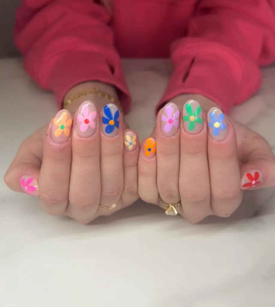 Hands with each nail painted with a different color base and adorned with simple, colorful flower designs, creating a cheerful springtime look.