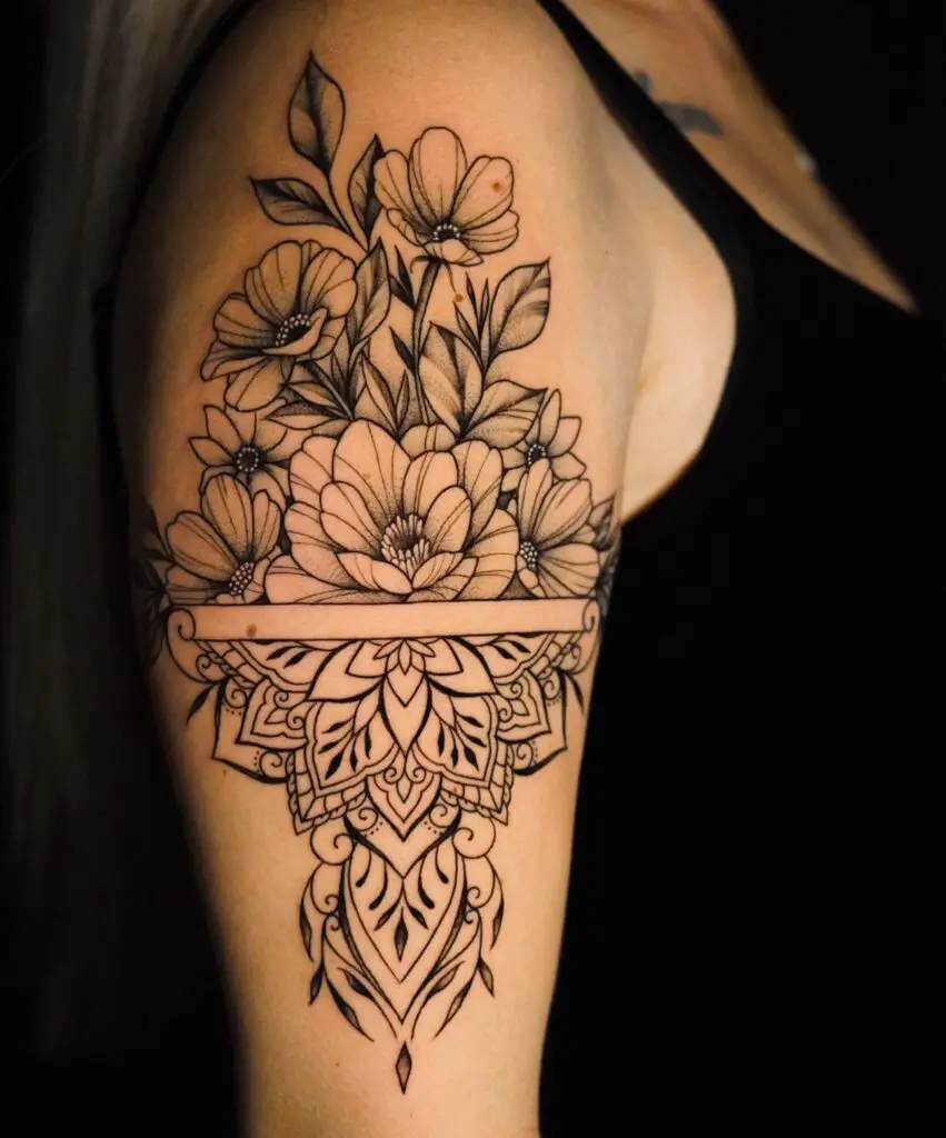 A black ink floral tattoo on an upper arm, featuring a symmetrical design of flowers and leaves with a mandala center, highlighted by fine lines and dot shading.