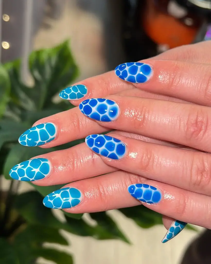 Stiletto nails featuring a blue honeycomb pattern with a gradient effect, set against a backdrop of lush green leaves.