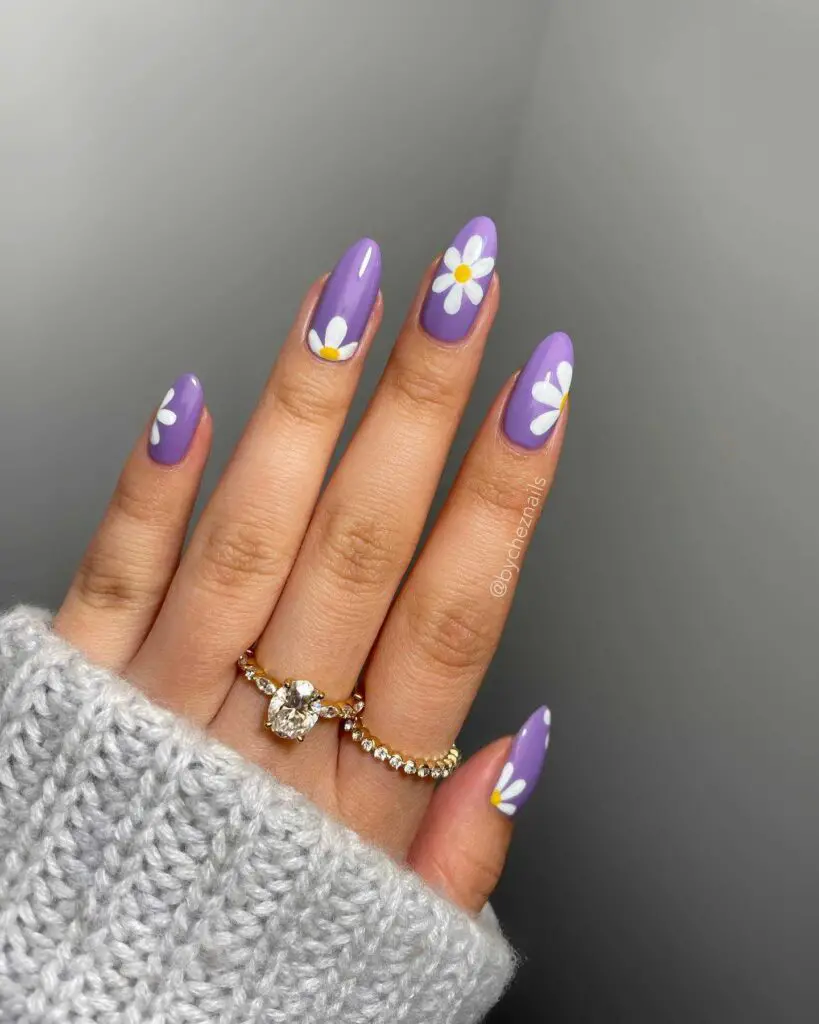 A hand with almond-shaped nails painted purple and decorated with white daisy designs, exemplifying a playful and stylish springtime manicure.