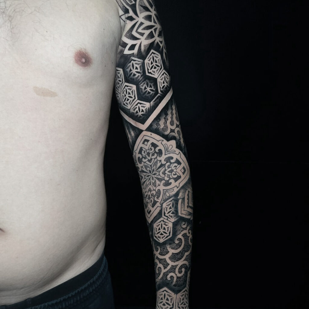 A full sleeve tattoo with a mix of geometric and traditional patterns, executed in black ink with varying shades and textures.