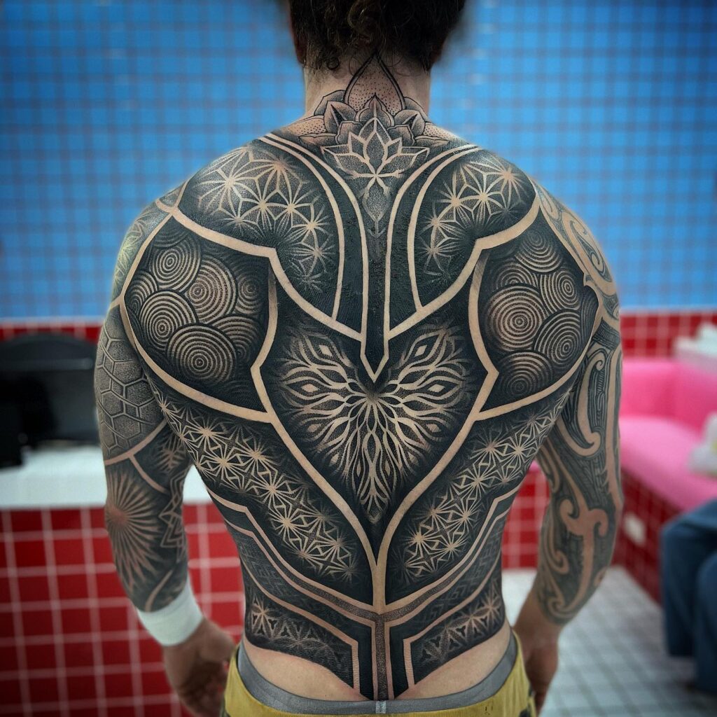 A detailed back tattoo with a central mandala, symmetric floral designs, and geometric patterns, inked in shades of black and gray.