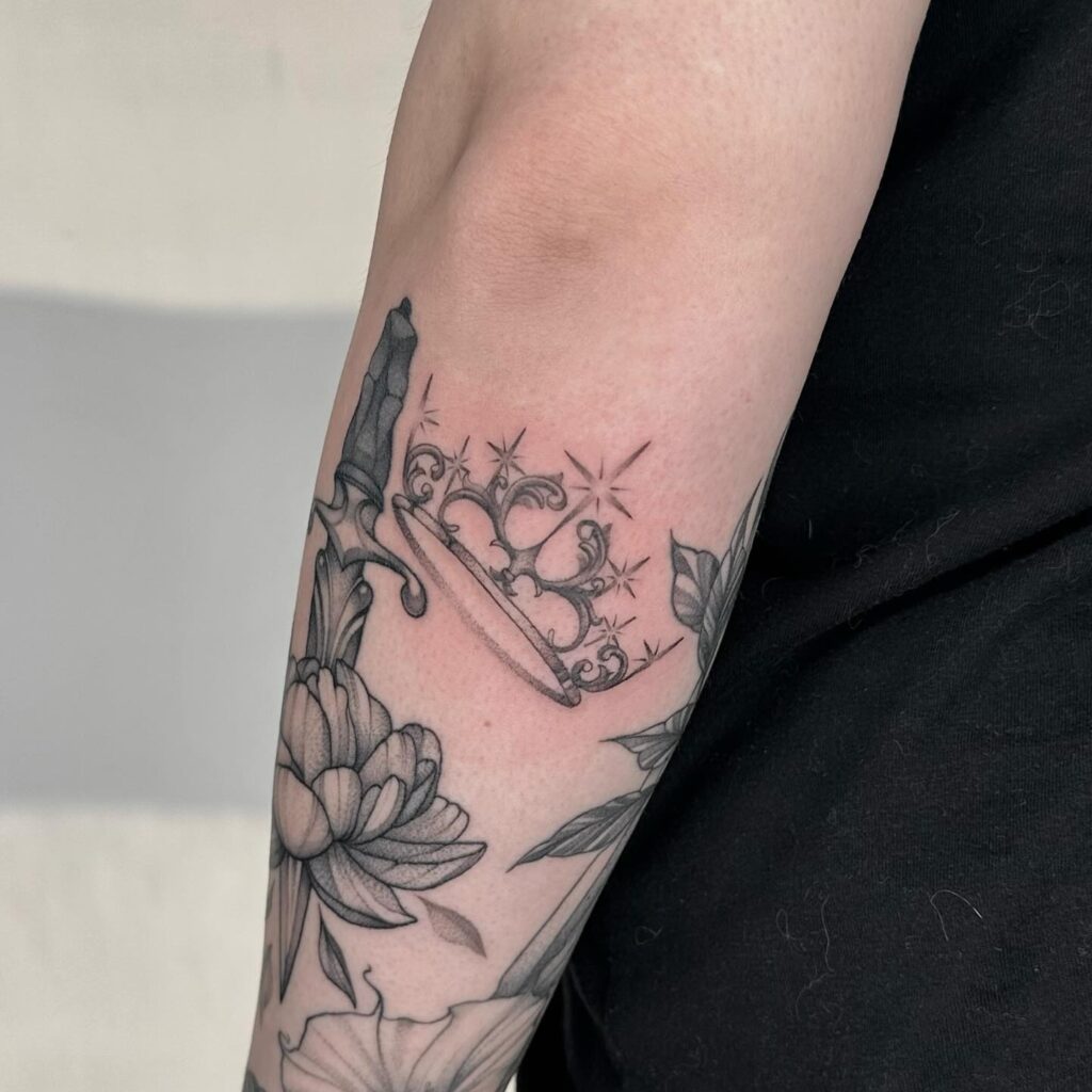 An arm tattoo depicting a sword with flowers through a crown, symbolizing the victory of beauty and intellect over might.