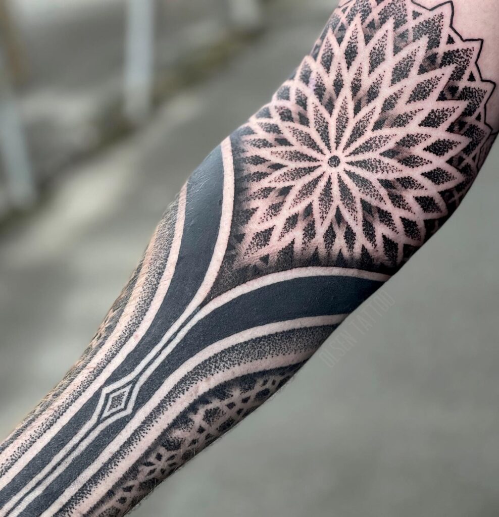 Forearm sleeve tattoo featuring a large elbow mandala surrounded by shaded geometric patterns and detailed dotwork.