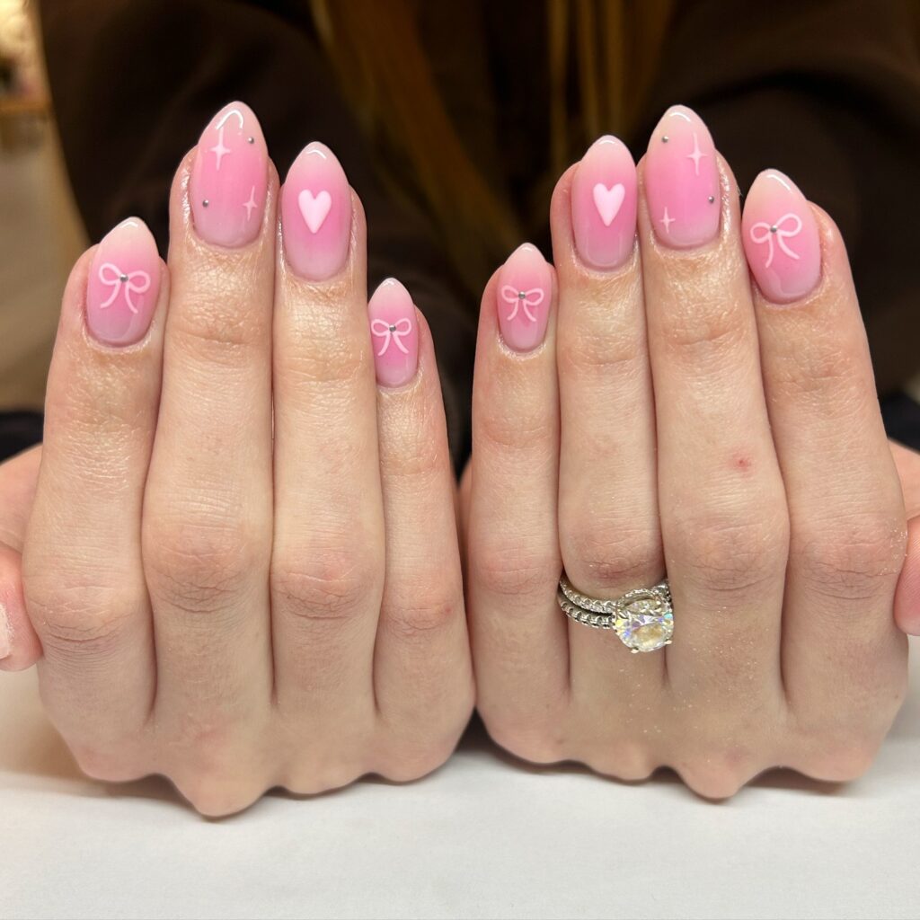 Hands with almond-shaped nails painted in soft pink, featuring cute hearts and bows for a lovingly crafted look.