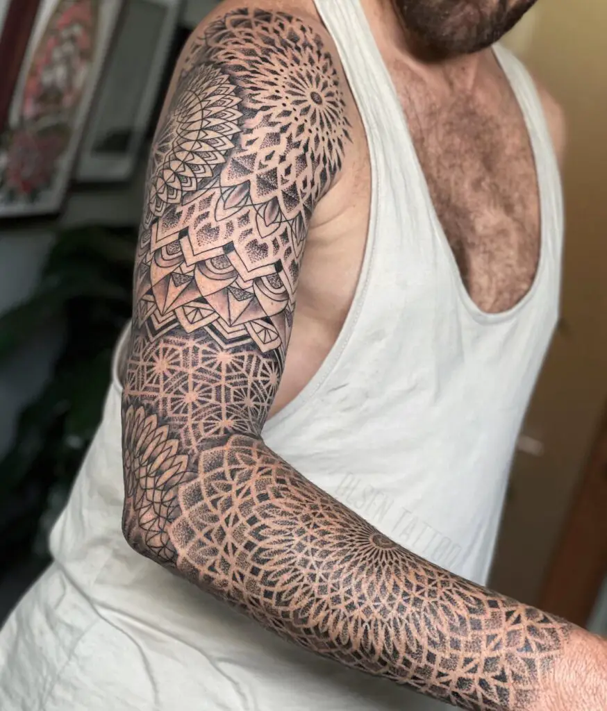 An upper arm tattoo blending a floral mandala with a sunburst geometric pattern, ending in a tight geometric mesh at the wrist.