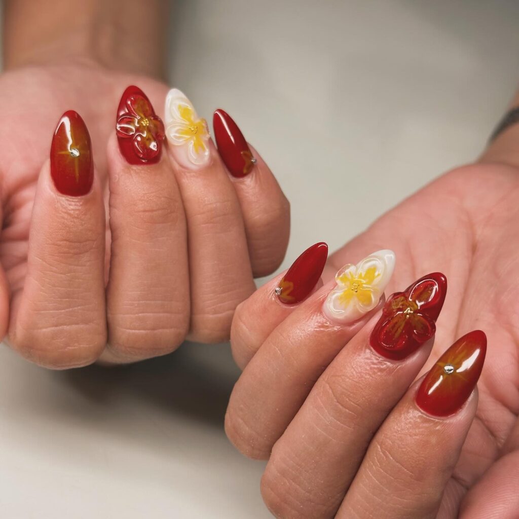 A pair of hands with glossy red nails, two of which are adorned with detailed 3D flower designs that add a touch of elegance and depth.