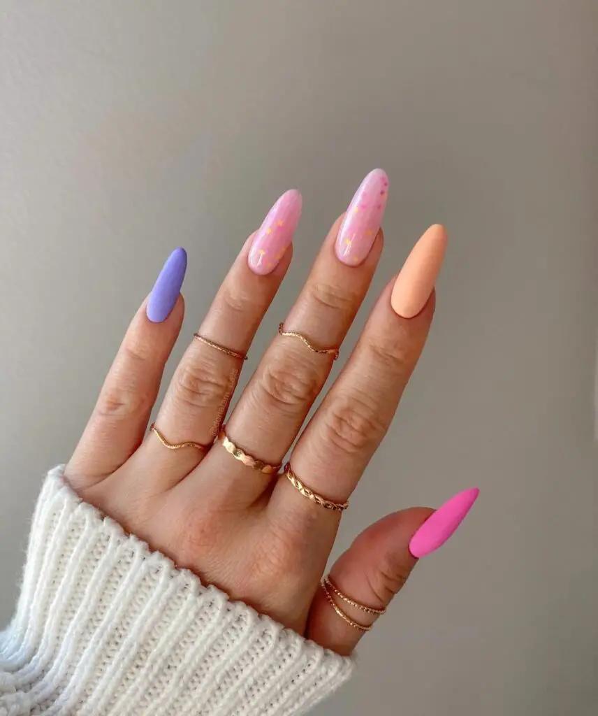 A hand showing nails painted in pastel shades of lilac and pink, with a striking matte peach on the thumb, all complemented by subtle glitter and star details.