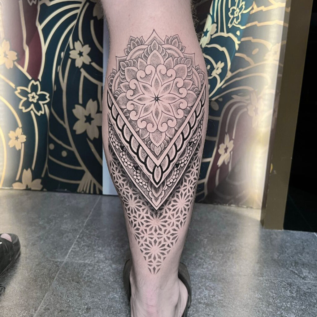 A detailed and layered mandala tattoo on a person's calf with a central floral design and geometric accents.