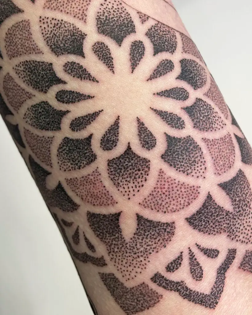 A close-up of a symmetrical mandala tattoo on a thigh, fading from black to pink with petal shapes and dotted details.